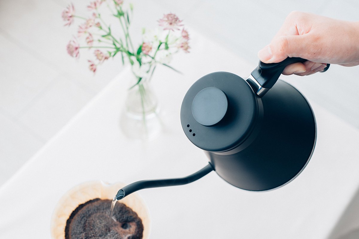 Person pouring coffee from a black kettle into a coffee filter on a white surface with flowers in the background.