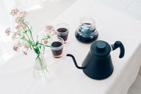 Black kettle, glasses of coffee, and a vase with flowers on a white surface
